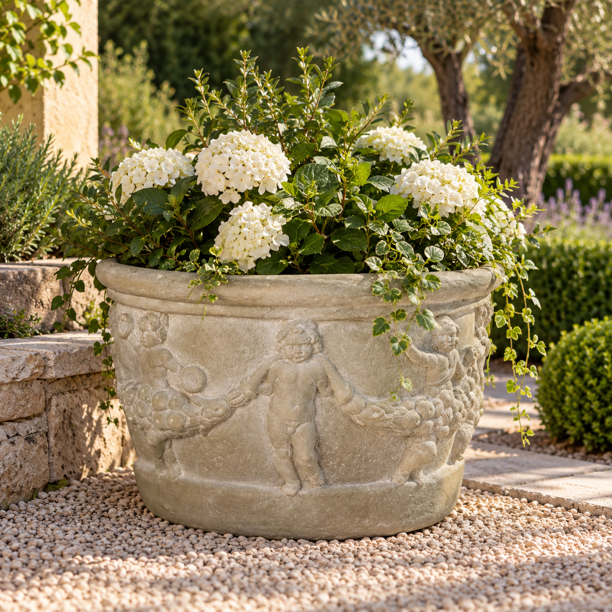 A large round stone planter with carved cherub figures, filled with white flowers and green foliage, placed outdoors on a gravel path in a garden setting.