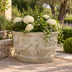 A large round stone planter with carved cherub figures, filled with white flowers and green foliage, placed outdoors on a gravel path in a garden setting.