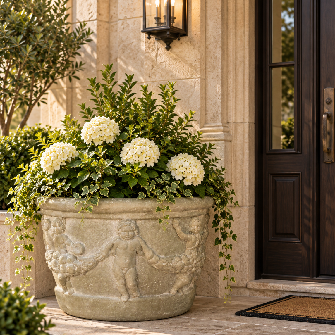 A large round stone planter with classical cherub carvings, filled with green foliage and white hydrangea blooms, placed on a stone porch beside a dark wooden door.