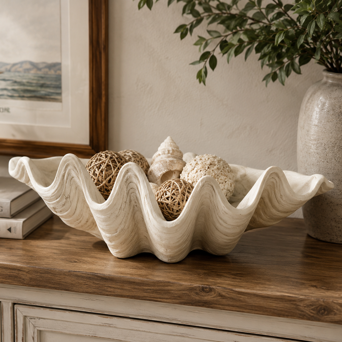 Decorative shell bowl with seashells on a wooden surface, with a framed picture and plant in the background.