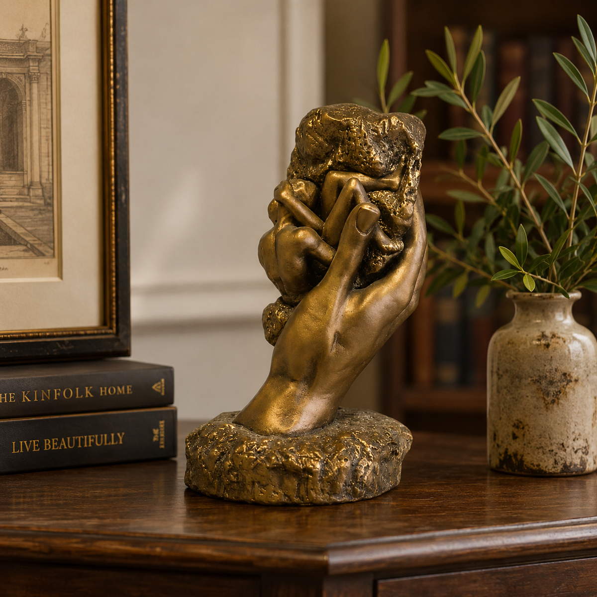 Bronze sculpture of a hand holding a head on a wooden surface with books and a vase in the background.