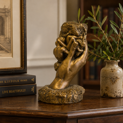 Bronze sculpture of a hand holding a head on a wooden surface with books and a vase in the background.