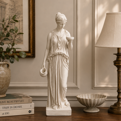 Statue of a classical woman holding a bowl on a wooden surface with books and decor items.