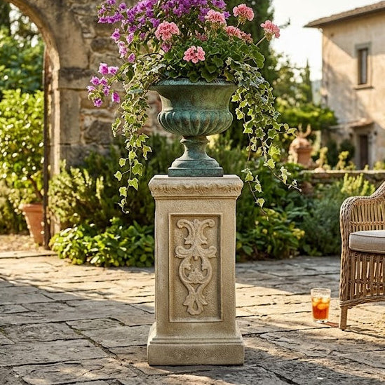 A large green stone urn planter overflowing with pink and purple flowers, displayed on a carved stone pedestal in a sunny, garden-chic courtyard next to a wicker armchair.
