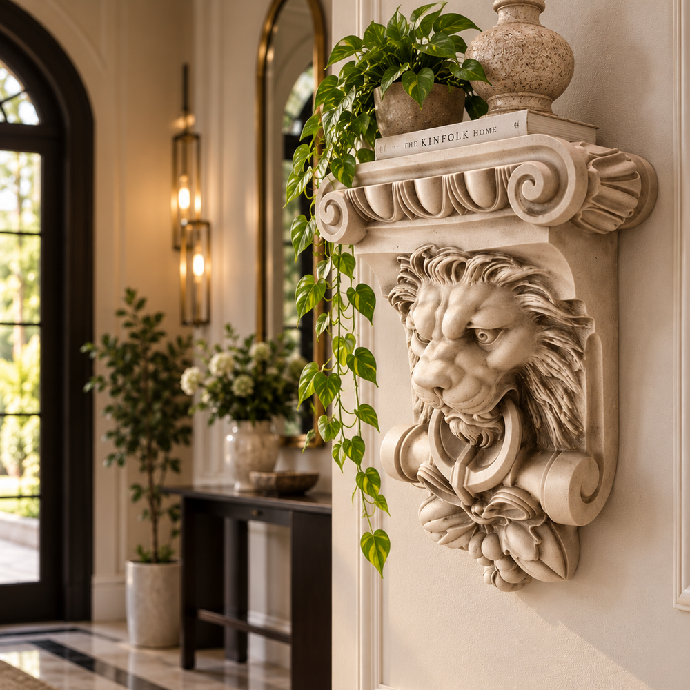 A decorative stone lion-head wall bracket mounted on a cream interior wall, styled with potted greenery and a vase. The background shows a softly blurred entryway with a table, sconces, mirror, and arched doorway.