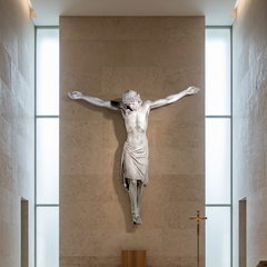 A white stone sculpture of Jesus Christ crucified, mounted on a large beige stone wall in a modern chapel or church interior with tall windows and a brass cross stand below.