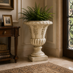 An ornate cream-colored stone-look urn planter filled with green ferns, placed in a classic entryway next to a dark wood console table and near a doorway.