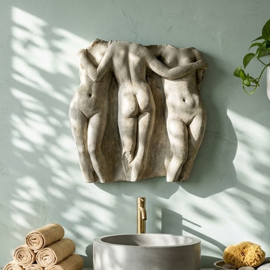 A styled bathroom interior with a pale green wall, a stone basin sink, rolled towels, a sponge, and a sculptural wall art piece depicting three intertwined figures. A clawfoot tub, hanging plants, and natural window light are visible in the room.