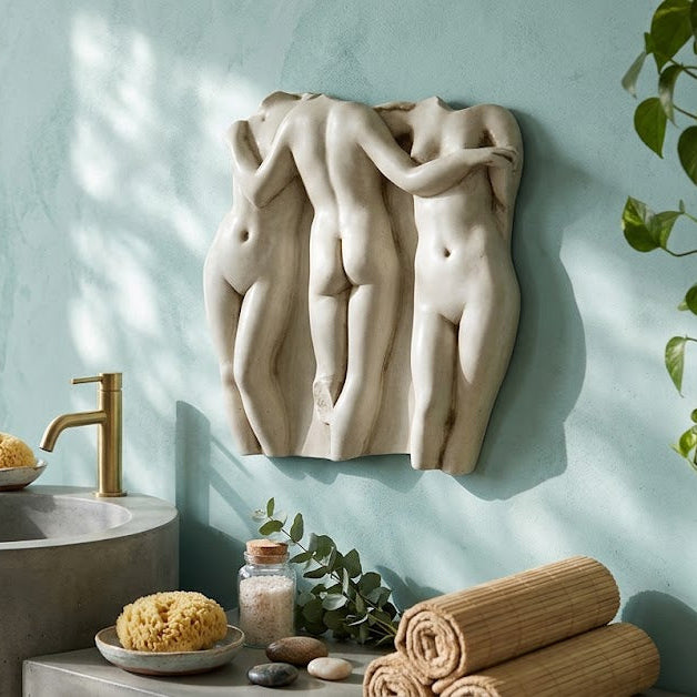 A decorative Three Graces Frieze 15  Wall Decor of three intertwined nude female figures in a light beige stone or plaster finish, mounted on a textured light blue wall in a styled bathroom setting with a concrete sink, gold faucet, towels, and plants.