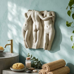 A decorative Three Graces Frieze 15  Wall Decor of three intertwined nude female figures in a light beige stone or plaster finish, mounted on a textured light blue wall in a styled bathroom setting with a concrete sink, gold faucet, towels, and plants.