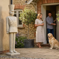 Couple with a dog standing outside a house with a garden statue and plants.