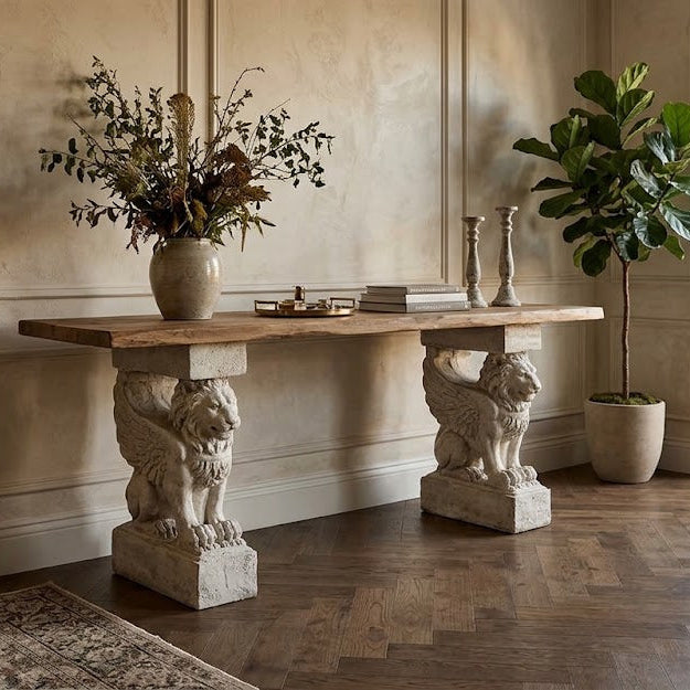 Winged Lion Consol Base - A rustic console table with a light wood top and two lion-carved stone legs, styled in a warm, elegant room with a window, potted plant, and armchair.