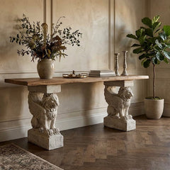 Winged Lion Consol Base - A rustic console table with a light wood top and two lion-carved stone legs, styled in a warm, elegant room with a window, potted plant, and armchair.