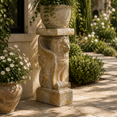 A stone lion pedestal planter holding a large green potted plant, set in an outdoor garden courtyard with stone paving, shrubs, and climbing vines.