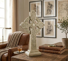 Decorative Celtic cross on a wooden table with books and a glass of whiskey, in a room with framed artwork on the wall.