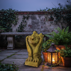 Gargoyle statue and lantern on a stone patio with plants and a stone wall in the background.