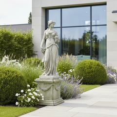 A classical-style stone statue of a draped female figure standing on a decorative pedestal, placed in a landscaped garden pathway with trimmed hedges, flowers, and a modern building with large glass windows in the background.
