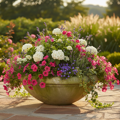 A large round stone planter filled with a colorful flower arrangement, including pink, white, and purple blossoms, set on a stone patio in a lush garden.