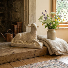 Decorative scene with a stone sculpture of a sheep, a vase of flowers, and a blanket on a wooden surface.